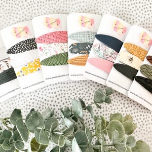 May include: A collection of hair clips in various patterns and colors, including floral, polka dot, and geometric designs. Each clip is displayed on a white card with the text "MYFRIENDSARAH". The clips are arranged on a dotted surface with greenery.