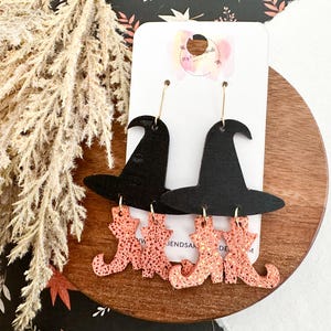 Peut inclure: Une paire de boucles d'oreilles sur le thème d'Halloween. Chaque boucle d'oreille présente un chapeau de sorcière noir et une paire de bottes de sorcière orange pailletées. Les boucles d'oreilles sont suspendues à des crochets dorés.