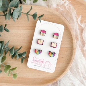 May include: Three pairs of stud earrings on a white display card labeled My Friend Sarah Made It. The designs include a stack of pink books, an open book with beige pages, and a heart-shaped arrangement of colorful books in rainbow hues.