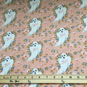 May include: Pink fabric with a repeating pattern of white ghosts with the word "BOO" printed on them. The ghosts are surrounded by small yellow and orange flowers. A wooden ruler is placed on the fabric.