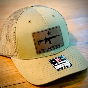 May include: A khaki green trucker hat with a brown mesh back. The hat features a black leather patch with a silhouette of a rifle and the text "Come and Take It". The hat also has a red and white label with the text "Richardson 115 Low Pro Trucker".
