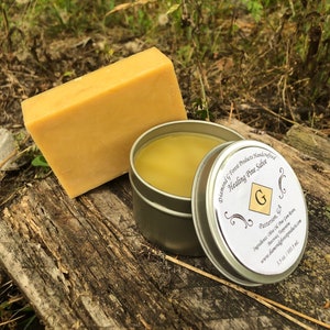 May include: A small tin of yellow healing pine salve with a label that reads "Diamond G Forest Products Handcrafted Healing Pine Salve" and "Patterson, GA". The tin is sitting on a piece of weathered wood with a bar of yellow soap behind it.