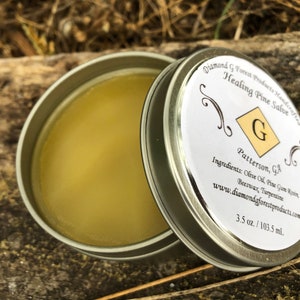 Puede incluir: Una lata de plata con una etiqueta blanca que dice "Diamond G Forest Products Handcrafted Healing Pine Salve" y "Patterson, GA". La etiqueta también enumera los ingredientes: aceite de oliva, resina de goma de pino, cera de abejas y trementina. La lata contiene un bálsamo amarillo. La lata es de 3,5 oz o 103,5 ml.