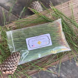 Può includere: Un sacchetto di plastica trasparente pieno di aghi di pino dell'Oregon verdi. Il sacchetto ha un'etichetta che recita "Diamond G's Slash Pine Needles" con una G dorata al centro.