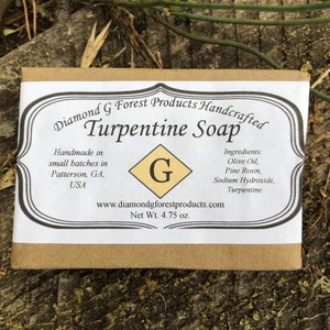May include: A brown paper wrapped bar of handmade "Turpentine Soap" by Diamond G Forest Products. The label is white with black text and a yellow diamond with a black "G" inside. The soap is made in small batches in Patterson, GA, USA and weighs 4.75 ounces. The ingredients are listed as olive oil, pine rosin, sodium hydroxide, and turpentine.