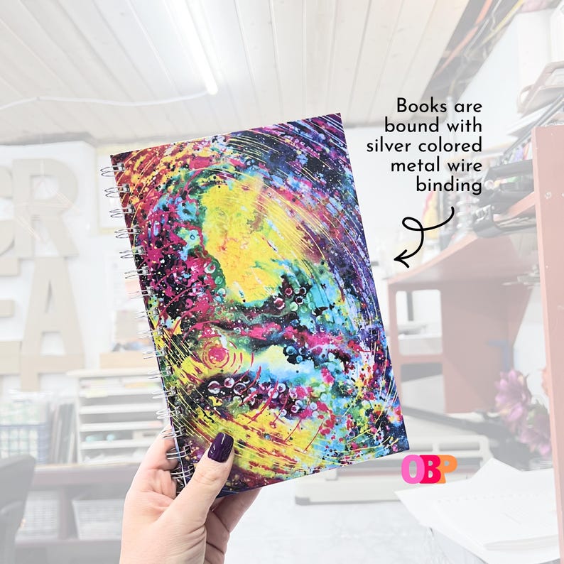 May include: A spiral-bound notebook with a vibrant, abstract design in yellow, pink, blue, and purple. The cover features a colorful, swirling pattern. The text "Books are bound with silver colored metal wire binding" is visible.
