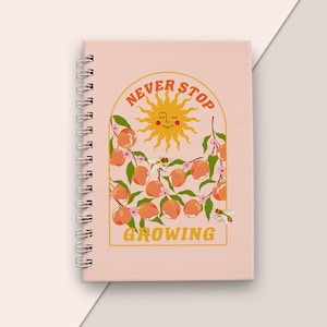 Pode incluir: Um caderno espiralado com capa rosa claro. A capa apresenta um design laranja com um sol sorridente, pêssegos e as palavras "Never Stop Growing". O design inclui abelhas e elementos florais.