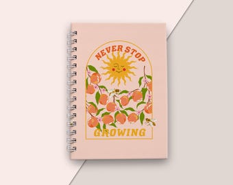May include: A spiral-bound notebook with a light pink cover. The cover features a design with a smiling sun, peaches, and the words "Never Stop Growing" in orange. The design includes bees and floral elements.