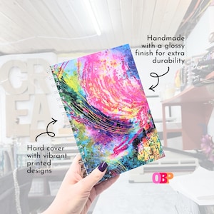 May include: A spiral-bound notebook with a hard cover featuring a vibrant, abstract design in pink, blue, yellow, and green. The cover has a glossy finish for durability. The text "Hard cover with vibrant printed designs" is visible.