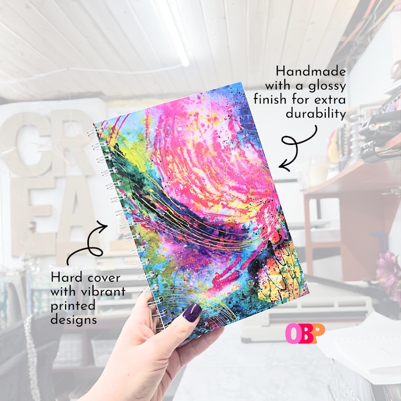 May include: A spiral-bound notebook with a hard cover featuring a vibrant, abstract design in pink, blue, yellow, and green. The cover has a glossy finish for durability. The text "Hard cover with vibrant printed designs" is visible.