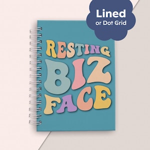Könnte beinhalten: Ein blaues Spiralnotizbuch mit dem Text "RESTING BIZ FACE" in bunten, Retro-Lettern.