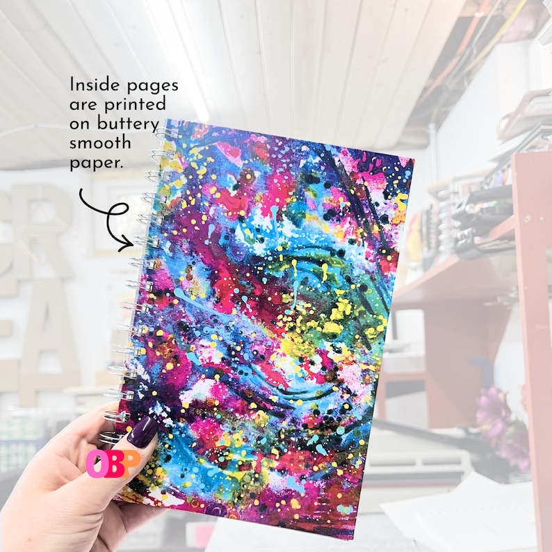 May include: A spiral-bound notebook with a vibrant abstract design. The cover features a colorful mix of pink, blue, yellow, and purple paint splatters. Text on the image reads, "Inside pages are printed on buttery smooth paper."