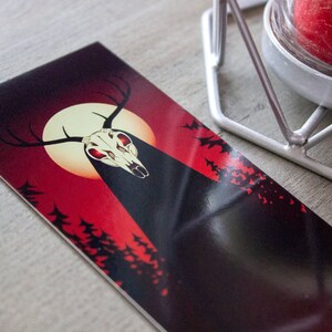 May include: A black and red illustration of a deer skull with glowing red eyes, set against a full moon and a dark forest background.