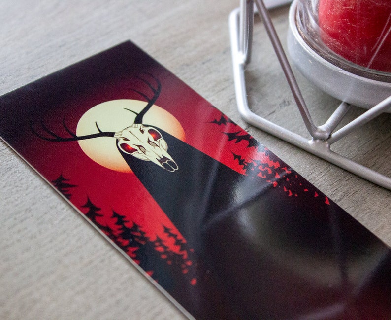 May include: A black and red illustration of a deer skull with glowing red eyes, set against a full moon and a dark forest background.
