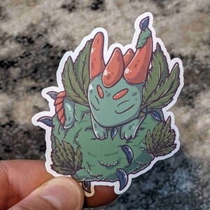 May include: A green and red cartoon creature with horns and leaves, sleeping on a green mossy mound.