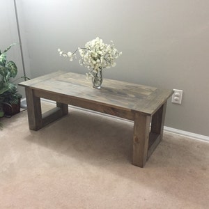 May include: A rustic wooden coffee table with a natural wood finish and a simple rectangular design. The table has a square base with open sides and sits on a beige carpet.