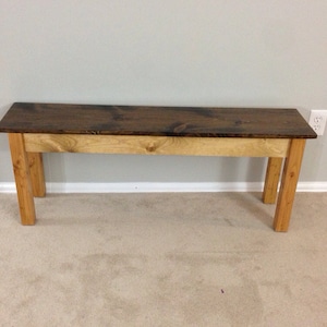 May include: A long, wooden bench with a dark brown stained top and natural wood legs. The bench is sitting on a light brown carpet.