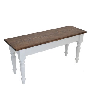 Puede incluir: Un banco de madera blanco con una tapa te&ntilde;ida de color marr&oacute;n oscuro y patas torneadas. El banco est&aacute; hecho de madera maciza y tiene un estilo sencillo de granja.