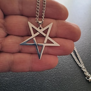 May include: A silver-colored pendant necklace featuring a pentagram design. The pendant is a five-pointed star with an inverted triangle in the center. The necklace has a chain with a lobster clasp. The background is a hand.