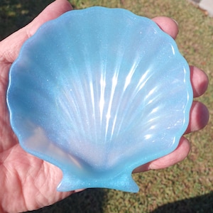 Può includere: Un portagioie a forma di conchiglia azzurra. Il piatto ha una finitura perlata con un motivo a ventaglio che si irradia dal centro. Il piatto è tenuto in mano, con uno sfondo verde sfocato.