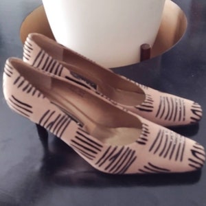 May include: A pair of blush-colored pumps with a black linear pattern. The shoes have a square toe and a small black heel. The shoes are on a dark surface, with a white pot of greenery in the background.