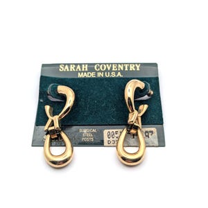 May include: Gold-tone Sarah Coventry earrings with a unique looped design. The earrings are displayed on their original green and black card, which reads "SARAH COVENTRY MADE IN U.S.A." and "SURGICAL STEEL POSTS."