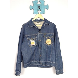 May include: A vintage denim jacket in a medium wash blue. The jacket features two chest pockets and two patches, one with flames and the other with a circular logo. It has a button-up front and a classic collar.