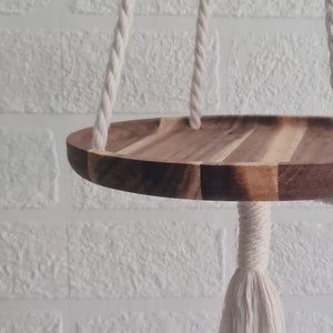 May include: A hanging wooden shelf with a round, segmented design. The shelf is suspended by thick, off-white rope and macrame. The background is a white brick wall. This shelf is perfect for displaying plants or decorative items.