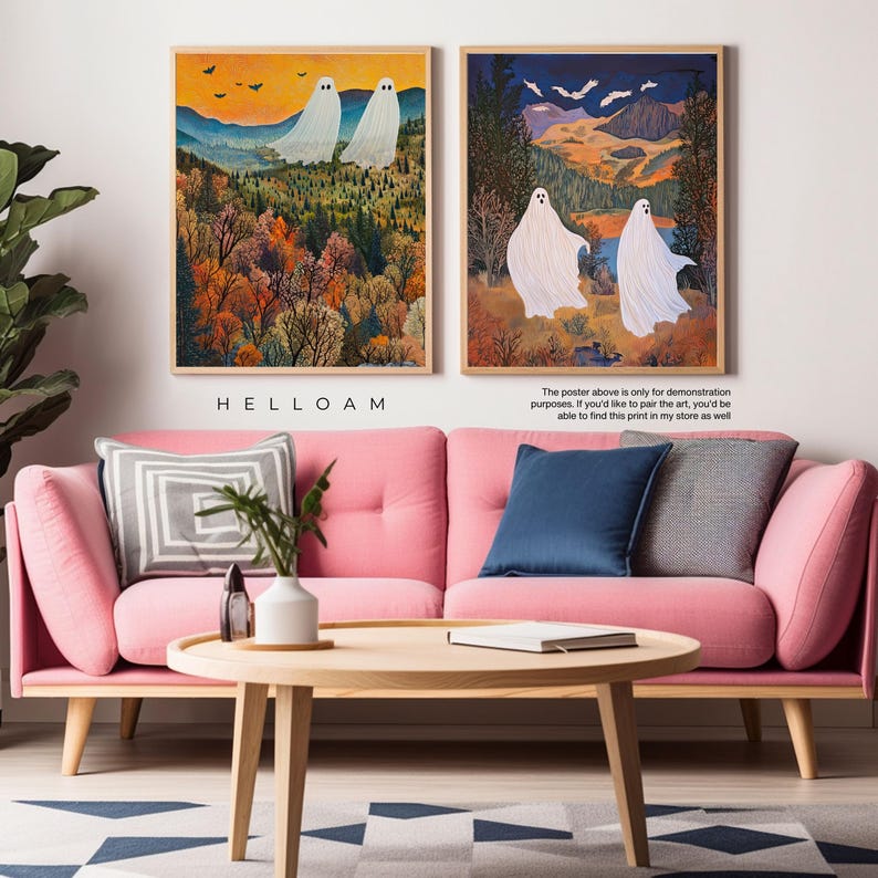 May include: Two digital art prints of white ghosts in a forest setting. The first print features two ghosts in a forest with fall foliage. The second print features two ghosts walking in a forest with a blue sky and mountains in the background. The prints are framed in wood and are displayed on a pink sofa with a coffee table.