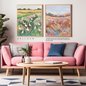 May include: Two framed paintings of landscapes with vibrant colors. The painting on the left features a field of flowers with white birds. The painting on the right features a field of tall grass with a mountain range in the background. The paintings are displayed on a pink sofa with pillows and a wooden coffee table.