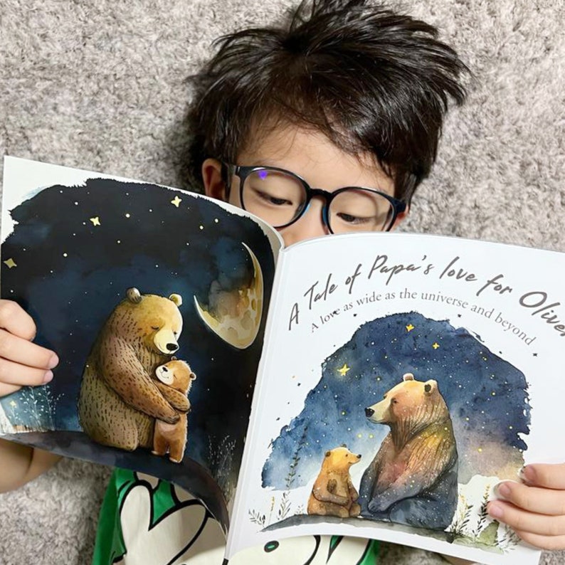 May include: A child reads a book titled "A Tale of Papa's Love for Olive" with a watercolor illustration of two bears on the cover. The book features a starry night sky with a crescent moon.