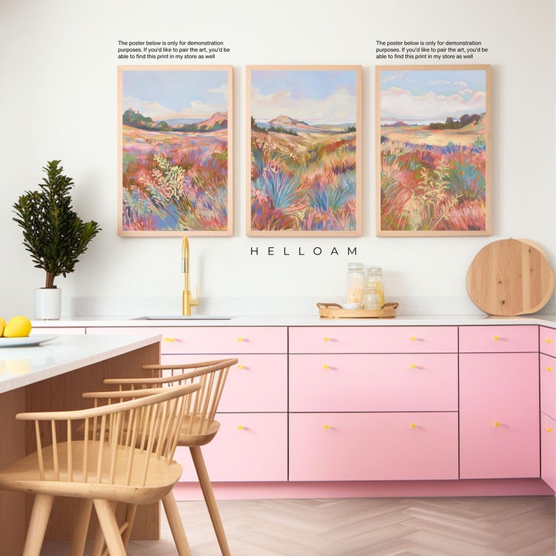 Peut inclure: Trois tableaux encadr&eacute;s de paysages dans un style aquarelle repr&eacute;sentant un champ d'herbes hautes et de fleurs dans des tons de rose, bleu et vert. Les tableaux sont accroch&eacute;s &agrave; un mur blanc au-dessus d'une armoire de cuisine rose avec des poign&eacute;es dor&eacute;es. Le texte "HELLOAM" est imprim&eacute; sous les tableaux.