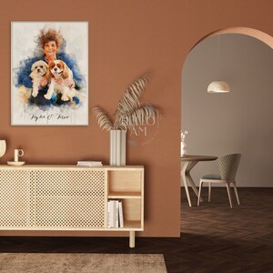 May include: A watercolor painting of a woman with two dogs. The woman is wearing a blue shirt and the dogs are brown and white. The painting is framed and hanging on a wall in a room with a wooden cabinet and a doorway.