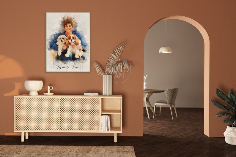 May include: A watercolor painting of a woman with two dogs. The woman is wearing a blue shirt and the dogs are brown and white. The painting is framed and hanging on a wall in a room with a wooden cabinet and a doorway.