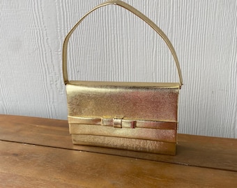 May include: A gold-colored clutch purse with a single top handle and a bow detail on the front.