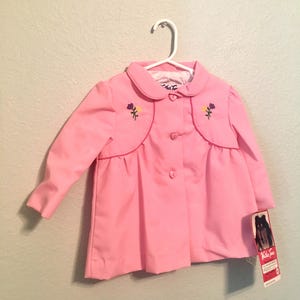Peut inclure: Une veste rose pour enfant avec un col Claudine et des fleurs brodées. La veste a une fermeture à boutons et un ourlet à volants. L'étiquette indique "Kids' Fashions".