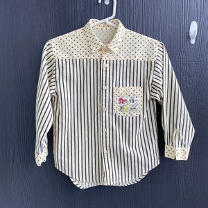May include: A white and black striped long-sleeved button-up shirt with a polka dot pattern on the top portion of the shirt. The shirt has a small pocket on the left side with a colourful embroidered design.