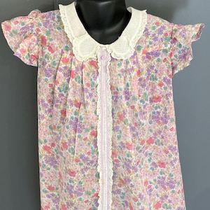 May include: A vintage-style floral print dress with a white lace collar and trim. The dress features a delicate pattern of pink, purple, and blue flowers on a light background. It has short sleeves and a button-up front.