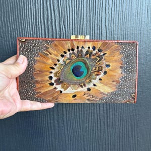 Könnte beinhalten: Rechteckige Clutch mit Pfauenfedermuster. Die Clutch hat einen braunen Lederrahmen und einen goldenen Verschluss. Die Mitte der Clutch zeigt ein lebendiges Pfauenauge mit Blau-, Grün- und Gelbtönen.