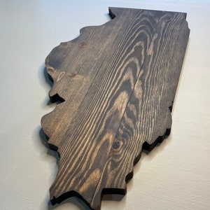 Puede incluir: Adorno de pared de madera con la forma del estado de Illinois. La madera tiene un tinte oscuro, resaltando la veta natural. Los bordes est&aacute;n cortados limpiamente y la pieza se muestra sobre un fondo neutro.
