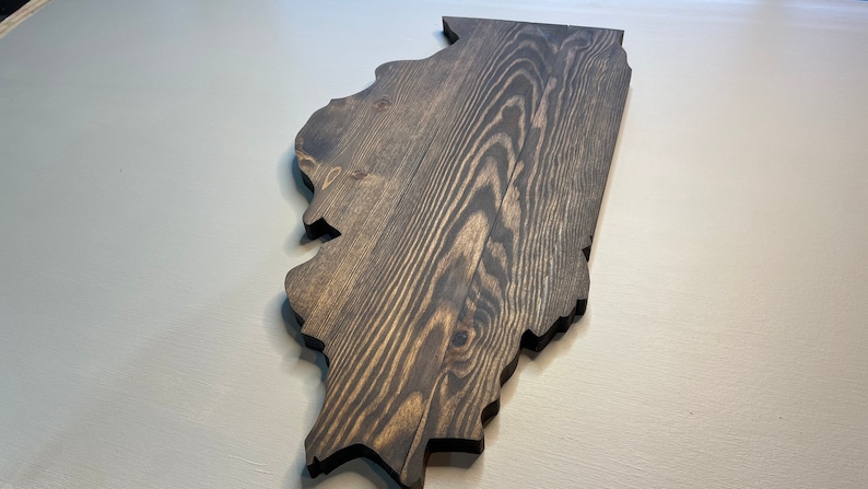 Puede incluir: Adorno de pared de madera con la forma del estado de Illinois. La madera tiene un tinte oscuro, resaltando la veta natural. Los bordes est&aacute;n cortados limpiamente y la pieza se muestra sobre un fondo neutro.