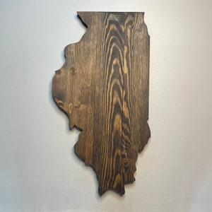 Puede incluir: Adorno de pared de madera con la forma del estado de Illinois. La madera tiene un tinte marr&oacute;n oscuro, con vetas visibles. Montado en una pared gris claro, resalta el contorno del estado. Mide aproximadamente 76 cm de alto.