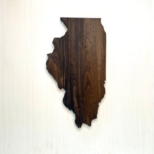 Puede incluir: Un recorte de madera con la forma del estado de Illinois. La madera tiene un tinte marr&oacute;n oscuro, resaltando la veta natural. El recorte se muestra sobre un fondo blanco.