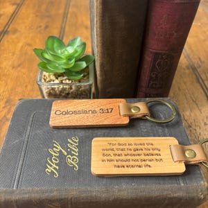 May include: Two wooden keychains with leather straps and metal rings. One keychain reads "Colossians 3:17", the other has a Bible verse. The keychains are on a Bible with the words "Holy Bible" in gold lettering.