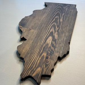 Puede incluir: Un recorte de madera del estado de Illinois, te&ntilde;ido en un color gris oscuro. La veta de la madera es visible, agregando textura a la pieza. Los bordes est&aacute;n cortados limpiamente y la pieza se muestra sobre una superficie gris claro.
