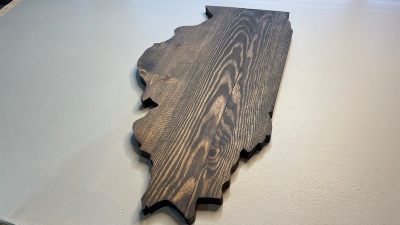 Puede incluir: Un recorte de madera del estado de Illinois, te&ntilde;ido en un color gris oscuro. La veta de la madera es visible, agregando textura a la pieza. Los bordes est&aacute;n cortados limpiamente y la pieza se muestra sobre una superficie gris claro.