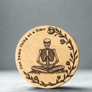 May include: A round wooden coaster with a laser-etched design. The design features a skeleton in a seated yoga pose, surrounded by floral elements. The text "One Damn Thing at a Time" is etched around the edge.