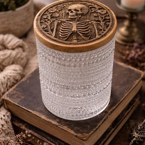 May include: A clear glass jar with a textured surface and a wooden lid. The lid features a carved design of a skeleton. The jar sits on top of two antique books. A candle in a brass holder is in the background.