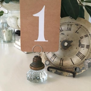 May include: A brown card with the number "1" printed in white is attached to a clear glass knob with a brass base. The knob is attached to a metal clock face.