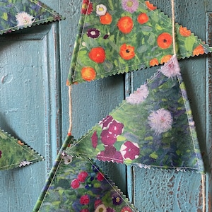 May include: A garland of triangular fabric bunting featuring a floral design in green, orange, white, and purple hues. The pennants are strung on natural twine, with white zigzag stitching along the edges. The backdrop is a weathered, light blue wooden surface.